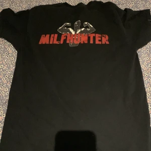Svart t-shirt med MILFHUNTER-tryck - Svart t-shirt med stort rött MILFHUNTER-tryck och en siluett av en muskulös person som flexar på bröstet. Klassisk passform och rund halsringning. Perfekt för dig som vill sticka ut med en bold statement-tee.