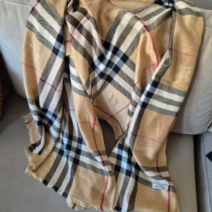 Rutig beige halsduk - Klassisk halsduk i 100% kashmir. Beige botten med rutigt mönster i svart, vitt och röda linjer. Mjuk och lyxig känsla, perfekt att slänga över axlarna för en snygg look. Tidlös design med fransar i kanterna.