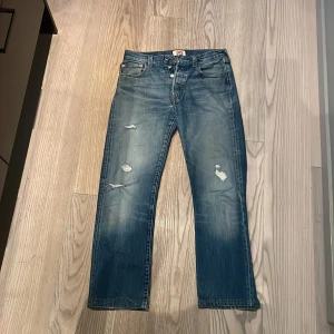 Levi's 501 blå jeans med slitningar - Klassiska Levi's 501 jeans i blå tvätt med snygga slitningar och hål på benen för en cool, avslappnad look. Rak passform, fem fickor och knappgylf. Perfekta för dig som gillar vintage vibe och tidlös denim.