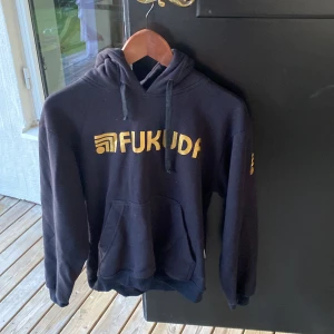 Svart hoodie från Fukuda med tryck - Svart hoodie från Fukuda med guldigt tryck på bröstet och ärmen. Klassisk känguruficka framtill, snörning i huvan och mjukt material. Perfekt för dig som gillar streetwear och vill ha en chill look.