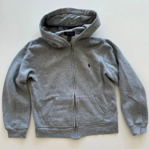 Grå hoodie från Polo Ralph Lauren - Klassisk grå hoodie från Polo Ralph Lauren med dragkedja framtill, huva och två fickor. Liten broderad logga på bröstet. Perfekt för en avslappnad stil och enkel att matcha med andra plagg.