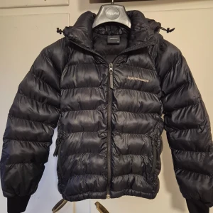Svart vinterjacka Peak Performance 140 - Svart vinterjacka från Peak Performance med broderad logga på bröstet och detalj på ärmen. Jackan har dragkedja framtill, två sidofickor med dragkedja och huva. Perfekt för kalla dagar och riktigt snygg streetstil.