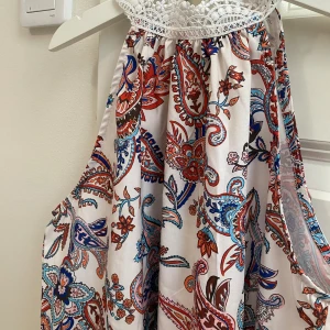 Vit topp med paisleymönster - Supersöt vit topp med färgglatt paisleymönster i blått, rött och orange. Toppen har en halterneck med vit spets upptill och är ärmlös. Materialet känns lätt och flowy, perfekt för varma dagar. Paisleymönstret ger en boho-vibe och klänningen har en loose passform💗