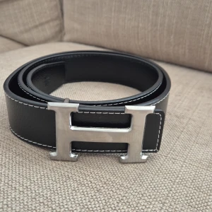 H-bälte - Snyggt bälte i svart skinn med ikoniskt,H-spänne i silverfärgad metall. Bältet har stilrena, kontrastsömmar och klassisk design som lyfter, vilken outfit som helst. Strl 110cm