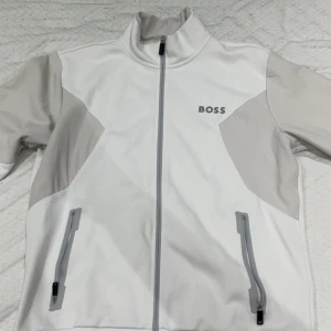 Vit vindjacka från BOSS, storlek L - Snygg vit vindjacka från BOSS med ljusgrå detaljer och hög krage. Jackan har hel dragkedja framtill, två fickor med dragkedja och logga på bröstet. Tillverkad i polyester för en sportig och clean look.