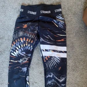 Stronger leggings med färgglatt mönster XS - Snygga leggings från Stronger i storlek XS med svart bas och färgstarkt abstrakt mönster i orange, vitt och blått. Bred resår i midjan med logga och två vita ränder på ena benet. Tillverkade i återvunnen polyester och elastan för en stretchig och bekväm passform.