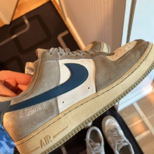 Nike Air Force 1 Club grå/blå sneakers - Nike Air Force 1 Club sneakers i grå mocka med vita paneler och blå swoosh. Klassisk låg modell med snörning, perforerad tåbox och beige sula. Snygg retrostil som passar perfekt till streetwear. Tydlig Air Force 1-brodyr på sidan och Club-märke på hälen. Köpt från fotolocker i Stockholm. Finns bara skor.