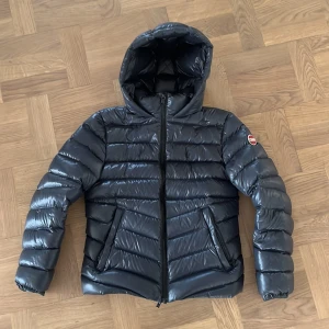 Svart Colmar pufferjacka med huva - Snygg svart pufferjacka från Colmar med glansig finish och huva. Jackan har dragkedja framtill, två fickor med dragkedja och klassisk Colmar-logga på ärmen. Perfekt för kalla dagar när du vill hålla stilen och värmen.
