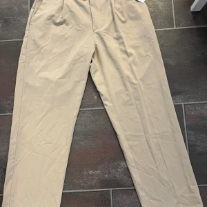 Beiga wide kostymbyxor från Bershka - Stilrena beiga kostymbyxor från Bershka i storlek L. Byxorna har en wide passform, hög midja, sidfickor och en bakficka. Tillverkade i ett mjukt bomullsmaterial med bälteshällor och knappstängning framtill. Perfekta för en clean och trendig look. Helt oanvända 