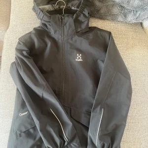 Svart vindjacka från Haglöfs - Säljer en svart vindjacka från Haglöfs med huva och dragkedja framtill. Jackan har reflekterande detaljer, meshfoder och är tillverkad i slitstark polyester. Perfekt för blåsiga dagar och outdoor-aktiviteter. Klassisk och stilren design med logga på bröstet.