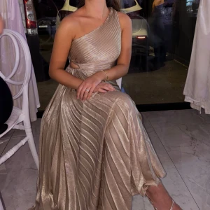 Champagnefärgad plisserad långklänning - Säljer en elegant champagnefärgad långklänning med plisserat tyg och metallicglans. Klänningen har oneshoulder-skärning och markerad midja med snygg cut-out på sidan. Perfekt för dig som vill sticka ut med en lyxig och trendig look.
