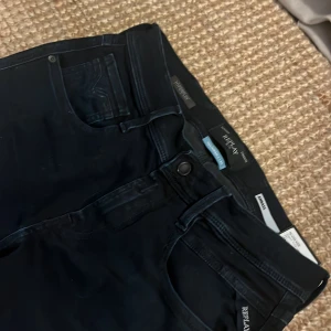 Svarta Replay Hyperflex jeans - Snygga svarta Replay Hyperflex jeans med klassisk femficksdesign och stretchigt material för extra komfort. Byxorna har bälteshällor och diskreta detaljer, perfekt för dig som gillar stilrena och moderna jeans.