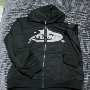 Svart hoodie med tryck och dragkedja - Svart hoodie med huva och hel dragkedja framtill. Stor tryckt logga med stjärndetaljer på bröstet. Mjukt material och ribbade muddar vid ärmslut och nederkant. Perfekt för en chill och avslappnad stil.