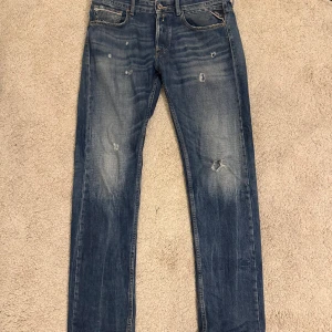 Replay Grover straight fit jeans blå - Säljer ett par Replay Grover jeans i straight fit med snyggt slitna detaljer och hål för en cool look. Färgen är klassiskt blå med ljusare tvätt och whiskers. Jeansen har fem fickor, knappgylf och är tillverkade i jeansmaterial. Perfekt för dig som gillar streetstyle.