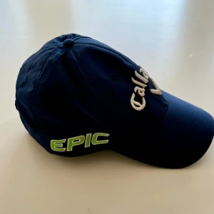 Mörkblå Callaway keps med brodyr - Snygg mörkblå keps från Callaway med broderade loggor och text i vitt, grönt och rött. Framsidan har Callaway-loggan, sidorna har EPIC, APEX och CHROME SOFT, och baktill står ODYSSEY med röd cirkel. Justerbar passform och klassisk böjd skärm. En riktigt snygg golf keps.