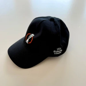 Svart UEFA Europa League keps - Svart keps med broderad UEFA Europa League-logga och trofésymbol i vitt och orange framtill. Justerbar metallspänne baktill för perfekt passform. Klassisk böjd skärm och ventilerande paneler. Perfekt för fotbollsfans som vill visa sitt stöd. fick den när jag var bollkalle på en uefa europa league match.
