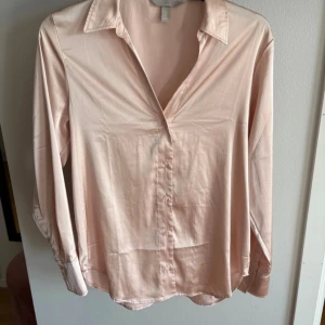 Ljusrosa skjorta från H&M - Säljer en stilren ljusrosa skjorta från H&M med klassisk krage och knäppning framtill. Skjortan har lång ärm och är tillverkad i ett mjukt, glansigt material som ger en snygg look. Perfekt att styla med jeans eller kjol.