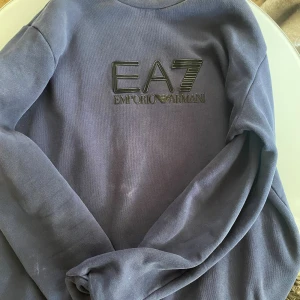 Mörkblå EA7 Emporio Armani tröja - Säljer en stilren mörkblå tröja från EA7 Emporio Armani med stor logga framtill. Tröjan har rund halsringning och långa ärmar, tillverkad i mjukt material som känns skönt mot huden. Perfekt för dig som gillar klassisk och sportig stil.