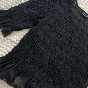 Svart volangblus från Gina Tricot - Supersnygg svart blus från Gina Tricot med volanger och rynkade detaljer över hela tröjan . Lätt och luftig med långärmade ärmar och transparenta partier. Perfekt för dig som vill sticka ut med en cool och trendig look.