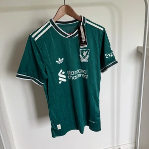 Grön Liverpool 25/26 fotbollströja Adidas M - Snygg grön Liverpool fotbollströja från Adidas med vita detaljer och tunna svarta ränder. Klassisk ribbad krage och ärmslut med vita och svarta inslag. Broderat klubbmärke och Adidas-logga på bröstet. Tillverkad i lätt och ventilerande material.