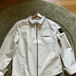 Beige overshirt från Stone Island - Snygg beige overshirt från Stone Island med klassisk krage, bröstficka och svart dragkedja framtill. Ikonisk patch på vänster ärm. Perfekt lager för vår och höst, stilren och enkel att matcha med det mesta.