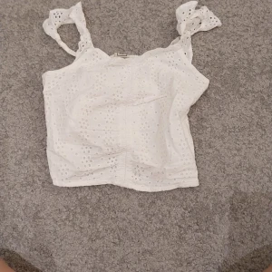 Vit broderad linnetopp från Ullared - Supersöt vit linnetopp med broderade hål och knytbara axelband. Toppen har smockad rygg för extra komfort och en luftig känsla. Perfekt för varma dagar och ger en romantisk vibe med sina volangdetaljer på axelbanden.