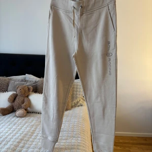 Beige mjukisbyxor Sail Racing M - Beiga mjukisbyxor från Sail Racing med broderad logga längs benet. Byxorna har snörning i midjan, muddar vid bensluten och är tillverkade i mjuk bomullsmix. Perfekta för chill eller träning, riktigt sköna och stilrena.