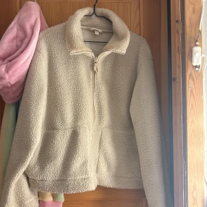 Beige teddyjacka från H&M, storlek L - Mysig teddyjacka från H&M i storlek L. Jackan är beige, har en hög krage och stängs med dragkedja framtill. Materialet är fluffigt och ger en chill vibe, perfekt för kalla dagar. Långärmad och med två fickor framtill för extra funktion.