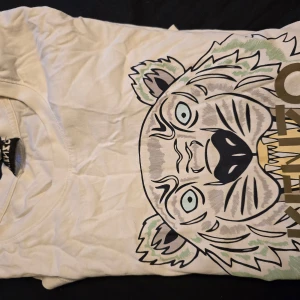 Vit Kenzo t-shirt med tigertryck - Vit t-shirt från Kenzo i bomull med stort, grafiskt tigertryck på framsidan i svart, grått, grönt och gult. Klassisk rund hals och korta ärmar. Perfekt statement-plagg för dig som gillar djärva prints och streetstyle.