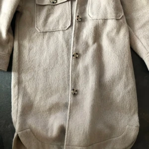 Beige overshirt från Gina Tricot - Säljer en beige overshirt från Gina Tricot med två stora bröstfickor och snygga knappar framtill. Jackan har en rak, loose passform och är tillverkad i mjukt ullmaterial. Perfekt att slänga över en hoodie för en chill look.
