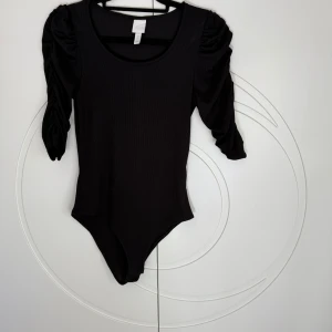 Svart ribbad body med puffärm - Säljer en svart ribbad body från H&M med snygga puffärmar och rund halsringning. Modellen har trekvartsärm och sitter tight mot kroppen. 