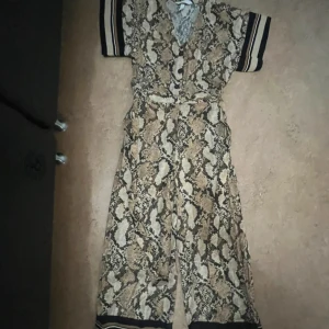 Ormmönstrad byxdress med knytband - Snygg byxdress med ormmönster i beige, brunt och svart. Vida ben och korta ärmar med svarta och vita ränder. Djup v-ringning framtill och knytband i midjan för extra passform. Perfekt för dig som vill sticka ut med ett coolt mönster.