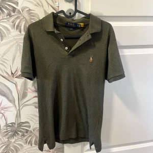Grå pikétröja Polo Ralph Lauren - Mörkgrå pikétröja från Polo Ralph Lauren i slim fit-modell. Klassisk krage, tre knappar och broderad logga på bröstet. Skön bomullskvalitet och korta ärmar, perfekt till jeans eller shorts.