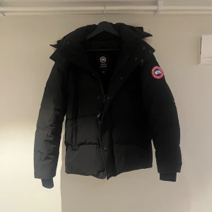 Svart dunjacka från Canada Goose - Säljer en svart dunjacka från Canada Goose med stor fluffig huva och beige pälsdetalj. Jackan har klassisk patch på ärmen, dragkedja och knappar framtill samt ribbade muddar vid ärmsluten. Perfekt för kalla vinterdagar och riktigt snygg passform.