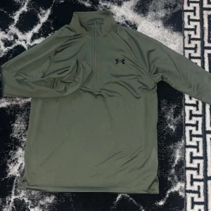 Mörk grön under armour tröja använd fåtal gånger.  Passar M/L  - Olivgrön långärmad tröja från Under Armour med half zip och hög krage. Tillverkad i mjukt och lätt polyestermaterial, perfekt för träning eller chill. Diskret svart logga på bröstet och nacken. Snygg och sportig stil som funkar året runt.