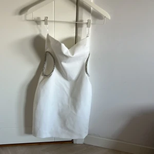 Vit kort klänning med strassdetaljer - Snygg vit kort klänning med tunna axelband och glittrande strassdetaljer runt utskurna sidor. Klänningen har en draperad urringning och figurnära passform, perfekt för dig som vill sticka ut. Dragkedja bak för enkel påklädning.