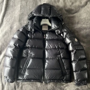 Moncler Maya - Säljer en svart pufferjacka från Moncler med glansig finish, huva och dragkedja framtill. Jackan har två fickor med dragkedja, en ärmficka med Moncler-logga och coolt serietryck på insidan. Perfekt för kalla dagar och riktigt snygg streetstil.