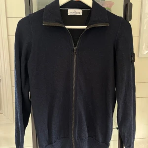 Mörkblå kofta från Stone Island Junior - Mörkblå kofta från Stone Island Junior med hel dragkedja framtill och klassisk ståkrage. Tröjan har lång ärm och ikonisk logotyp-patch på ena ärmen. Tillverkad i mjukt stickat material som passar perfekt till lager-på-lager-stil.