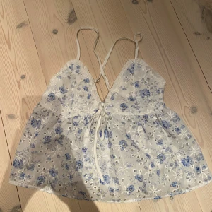 Blommig vit och blå linnetopp - Supersöt linnetopp med tunna axelband och blommigt mönster i vitt och blått. Toppen har broderade detaljer, v-ringning och snörning framtill. Perfekt för varma dagar och ger en romantisk vibe.