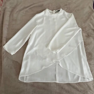Vit omlottblus från Zara - Säljer en vit omlottblus från Zara med lång ärm och hög hals. Blusen har en snygg omlott-detalj framtill och är tillverkad i ett lätt, flowy material som ger en elegant look. Perfekt för dig som gillar stilrena och trendiga plagg.