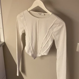 Vit ribbad croppad topp från NA-KD - Säljer en vit långärmad croppad topp från NA-KD i ribbad bomullsmix, storlek S. Toppen har coola sömdetaljer framtill och en figurnära passform som framhäver midjan. Perfekt till jeans eller kjol för en trendig look.