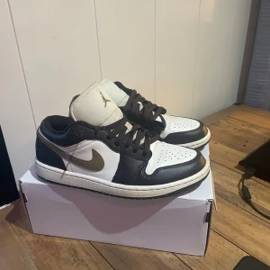 Nike Air Jordan 1 Low svart/vit - Snygga Nike Air Jordan 1 Low sneakers i svart och vitt skinn med bruna detaljer och klassisk Swoosh på sidan. Lågt skaft, svart snörning och perforerad tå för extra stilpoäng. Perfekta för dig som vill sticka ut med en ikonisk streetwear-look.