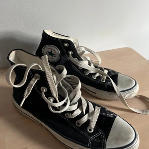 Mörkblåa Converse All Star high-tops - Klassiska marinblåa Converse All Star high-tops med vit sula och tå, vita snören och tydliga kontrastsömmar. Ikonisk logga på sidan och All Star-tryck bak på hälen. Skorna är i canvas och har rund tå samt platt sula.