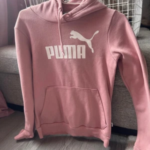 Rosa hoodie från Puma med logga - Säljer en rosa hoodie från Puma med stor vit logga framtill. Tröjan har huva med snörning, magficka och långa ärmar. Mjuk insida och klassisk passform, perfekt för chill eller streetstyle. Materialet är bomullsmix som känns skönt mot huden. Liten fläck under snöret se bild