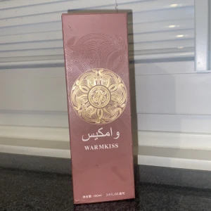Warmkiss parfym 100ml - Parfymen Warmkiss i en snygg rosabrun kartong med guldigt, orientaliskt mönster på framsidan. Flaskan rymmer 100 ml och har en lyxig känsla med sin eleganta förpackning. Perfekt för dig som gillar unika dofter och stilren design.