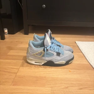 Nike Jordan 4 University Blue sneakers - Nike Jordan 4 University Blue sneakers med ljusblå mocka, svarta och vita detaljer samt stänkmönstrad häl. Klassisk siluett med meshpaneler, snörning och Jumpman-logga på baksidan. Perfekt för dig som vill sticka ut med en ikonisk sneaker.