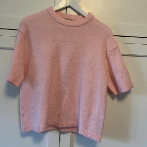 Ljusrosa stickad tröja med kort ärm - Superfin ljusrosa stickad tröja med korta ärmar och rund hals. Tröjan har ribbade muddar vid ärmslut och nederkant, vilket ger en snygg och enkel look. Perfekt för dig som gillar mjuka pasteller och en avslappnad stil.