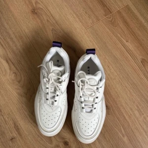 Vita sneakers från Eytys i skinn - Säljer ett par vita Eytys sneakers i skinn med chunky sula och lila hälflik. Skorna har perforerade detaljer på tån, snörning och en stilren design som passar till det mesta. Klassisk rund tå och platt sula för en clean look. Endast använda en gång. 