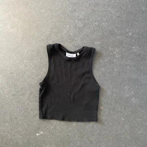 Svart croppad linnetopp Weekday - En svart, croppad linnetopp från Weekday med rund halsringning och ärmlös design. Toppen är i mjuk bomull och har en enkel, stilren look som passar perfekt till jeans eller kjol. Perfekt för varma dagar eller lager på lager.