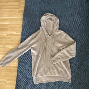 Beige Kashmir hoodie  - Mysig beige hoodie i stickat material med huva och långa ärmar. Tröjan har snörning med silvriga detaljer i ändarna och ribbade muddar vid ärmslut och nederkant. Perfekt för dig som gillar en chill och stilren look.